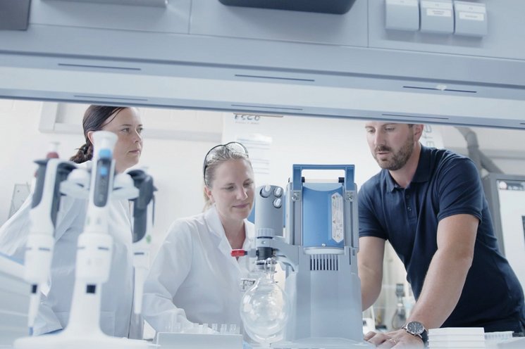 Two laboratory staff members and a technician look with satisfaction at a vacuum pump installed in a laboratory fume hood